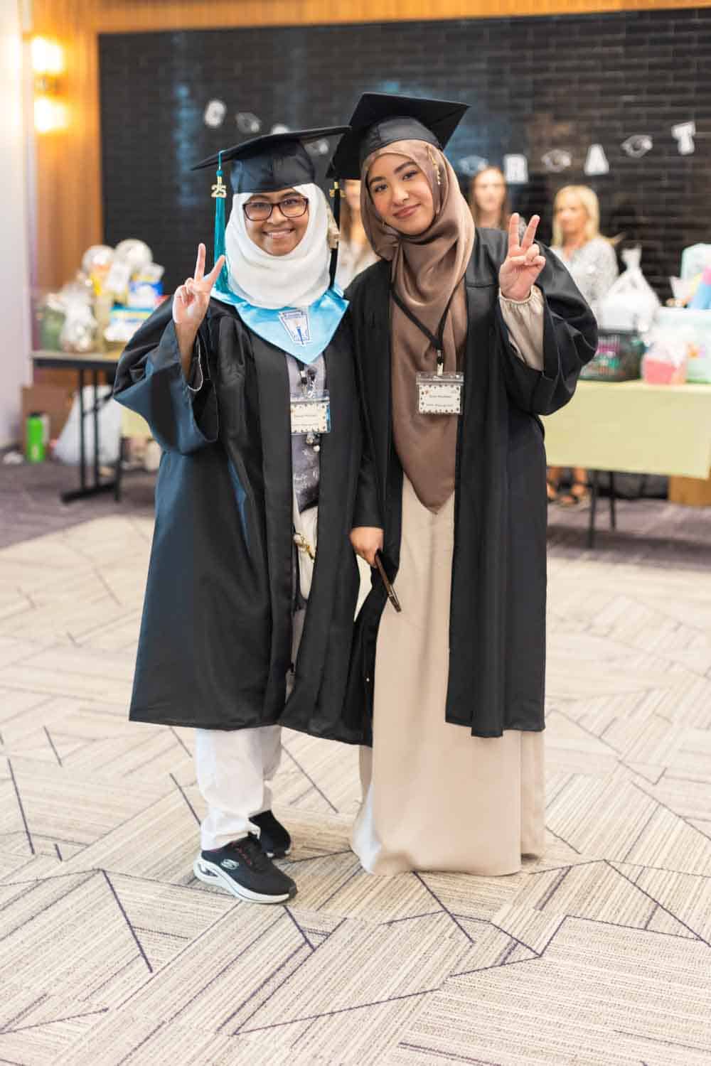 Two graduates in caps and gowns smile and pose with peace signs at a celebration. A festive backdrop and table with gifts are seen behind them.
