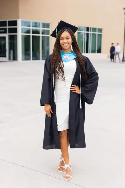 A young woman in a graduation cap and gown stands smiling outdoors on a sunny day, conveying a sense of achievement and joy.