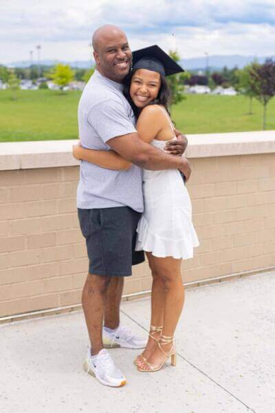 A proud father embraces his daughter in a graduation cap and white dress, both smiling warmly. They stand outdoors near a brick wall with a green field and mountains in the background.
