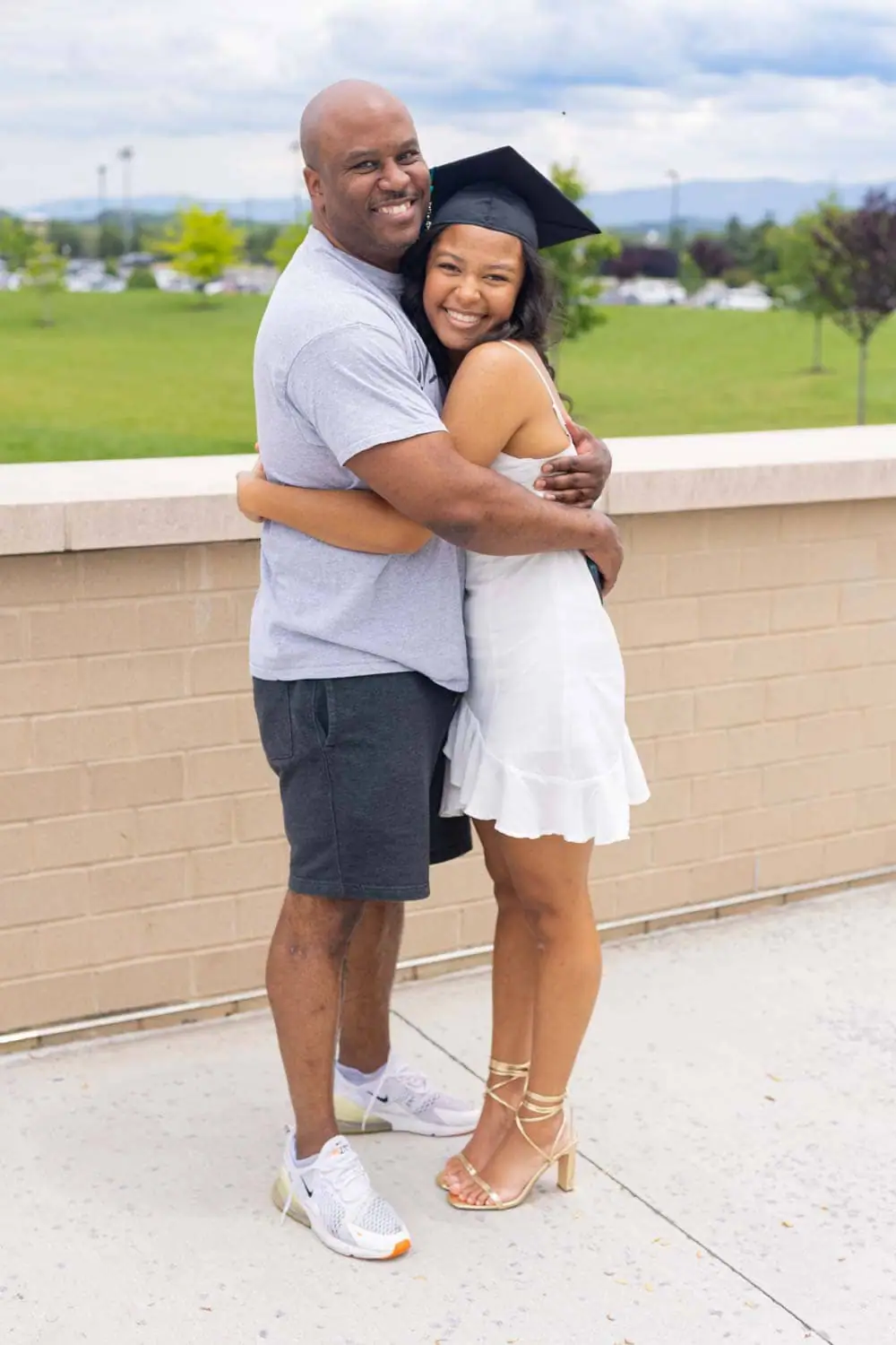 A proud father embraces his daughter in a graduation cap and white dress, both smiling warmly. They stand outdoors near a brick wall with a green field and mountains in the background.