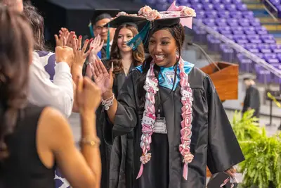 Graduating student in decorated cap and gown high-fives supporters, wearing a "2023" sash. Joyful atmosphere in an auditorium setting.