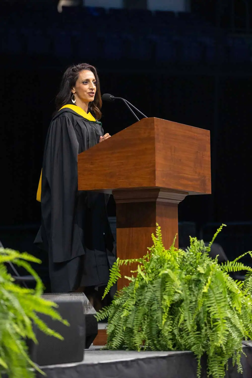 A woman in graduation robes speaks at a wooden podium on a stage. She appears confident as she addresses the audience. Green ferns are in the foreground.