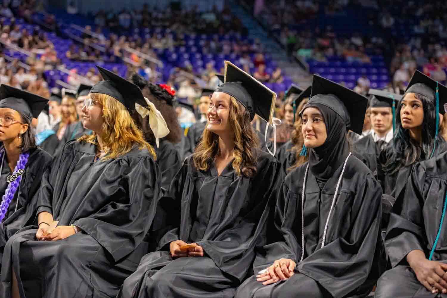 A group of graduates in black caps and gowns sit in a row at a graduation ceremony. They appear joyful and excited, with a packed audience in the background.
