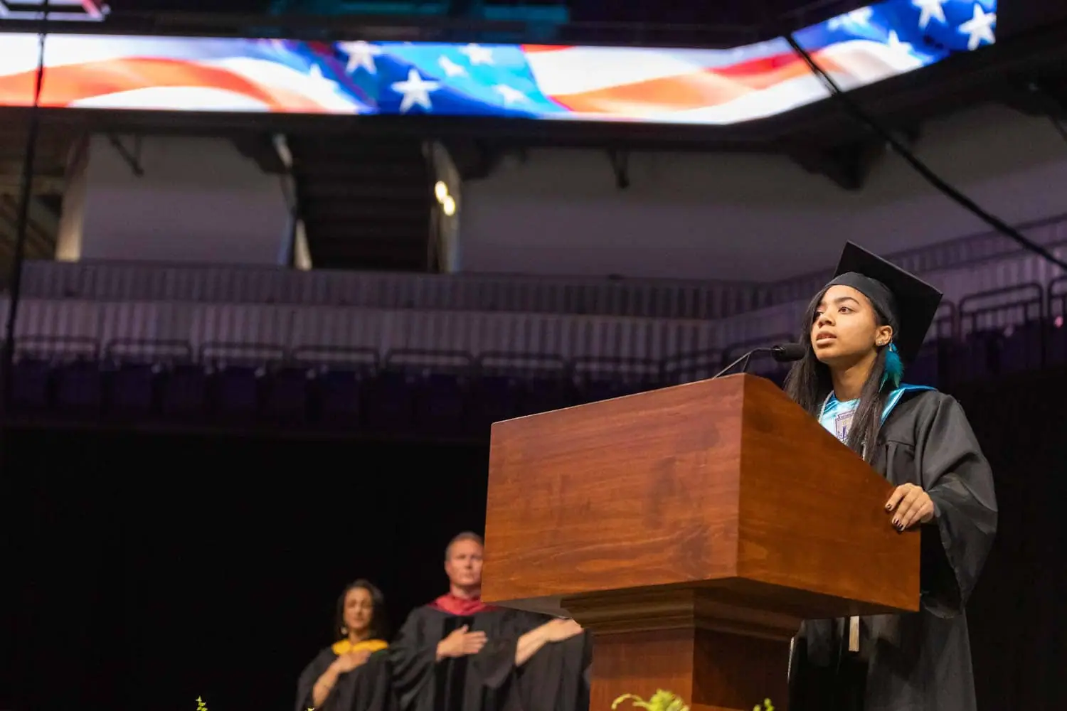 A graduate in a cap and gown speaks at a podium during a ceremony. The American flag is displayed on a screen above, with an audience in the background.