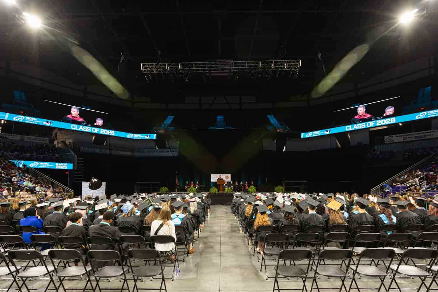 Wide-angle view of a graduation ceremony indoors. Graduates in caps and gowns face a stage with a podium. Screens display "Class of 2026."