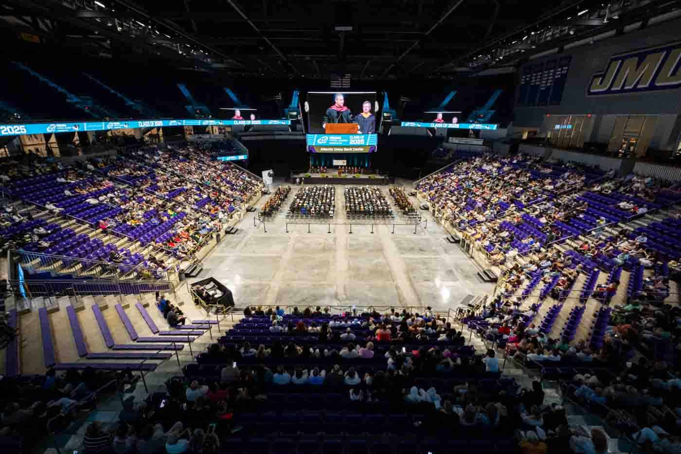 Large arena with purple seats filled with people attending a formal event. A stage with speakers is at the center, and a big screen displays them.