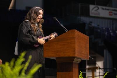 A woman in a graduation gown plays a ukulele at a wooden podium. She appears joyful, with a green plant in the foreground and a blurred auditorium background.