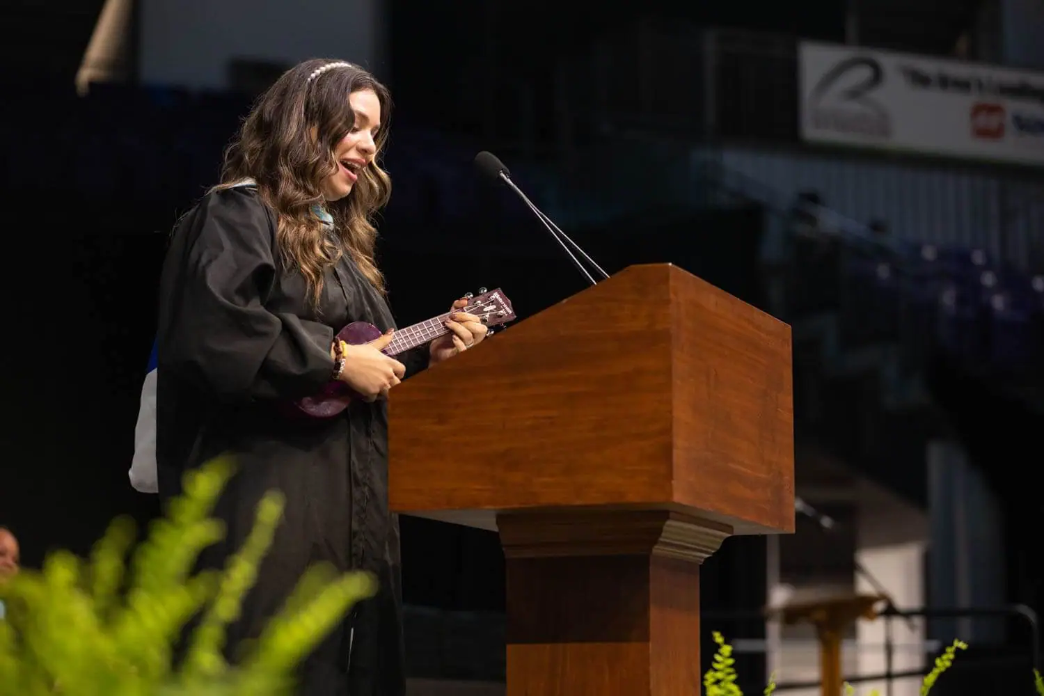 A woman in a graduation gown plays a ukulele at a wooden podium. She appears joyful, with a green plant in the foreground and a blurred auditorium background.