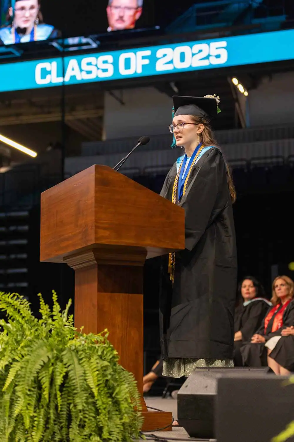 A graduate in cap and gown speaks at a podium labeled "Class of 2025" during a commencement ceremony. Audience members and ferns surround the stage.