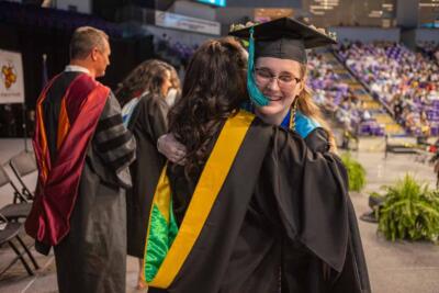 A graduate in a cap and gown smiles warmly while hugging another person in academic attire with a yellow sash. Other graduates and audience members are visible in the background.