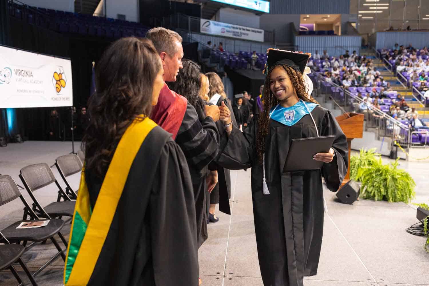 Graduation ceremony with a smiling graduate in cap and gown shaking hands with faculty on stage. Audience seated in background; cheerful and celebratory atmosphere.
