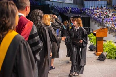 A graduate in cap and gown shakes hands with faculty on stage during a ceremony. Audience seated in the background, with a podium and plants nearby.