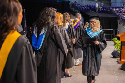 Graduate in cap and gown shakes hands with faculty in academic regalia on stage. The scene conveys achievement and celebration at a graduation ceremony.
