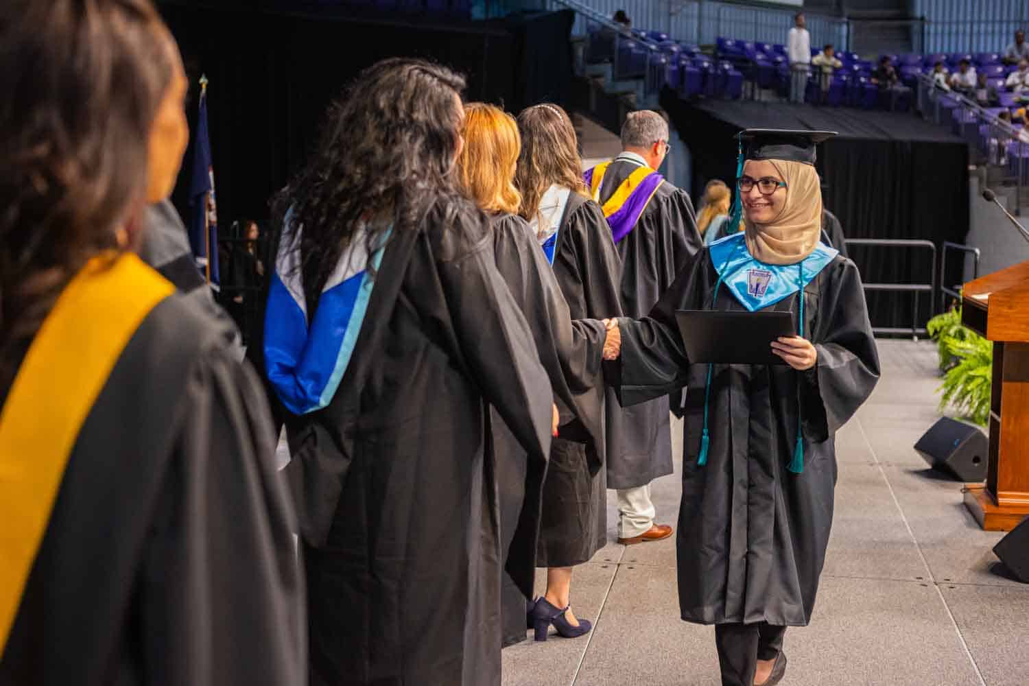 Graduate in cap and gown shakes hands with faculty in academic regalia on stage. The scene conveys achievement and celebration at a graduation ceremony.