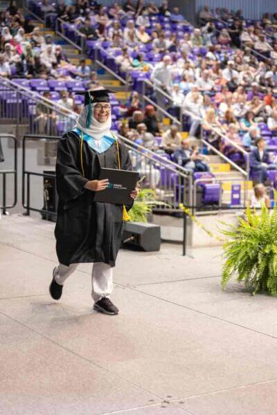 A graduate in a cap and gown joyfully walks across a stage with a diploma, surrounded by a cheering audience in a large auditorium setting.