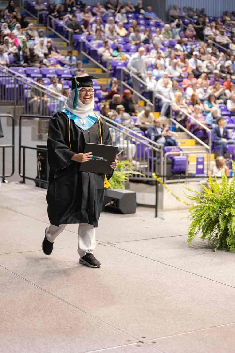 A graduate in a cap and gown joyfully walks across a stage with a diploma, surrounded by a cheering audience in a large auditorium setting.