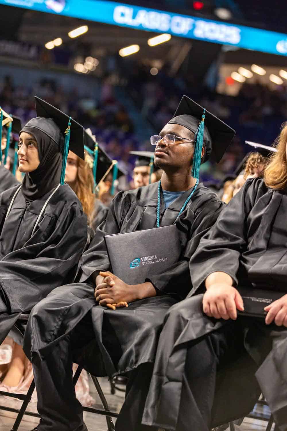 Graduates in caps and gowns sit attentively during a ceremony. One holds a diploma folder. The atmosphere is celebratory and focused.