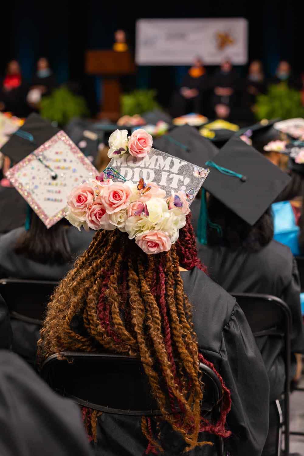 Graduation ceremony with attendees in caps and gowns. A decorated cap in focus reads "Mommy did it!" with pink and white flowers, conveying joy.