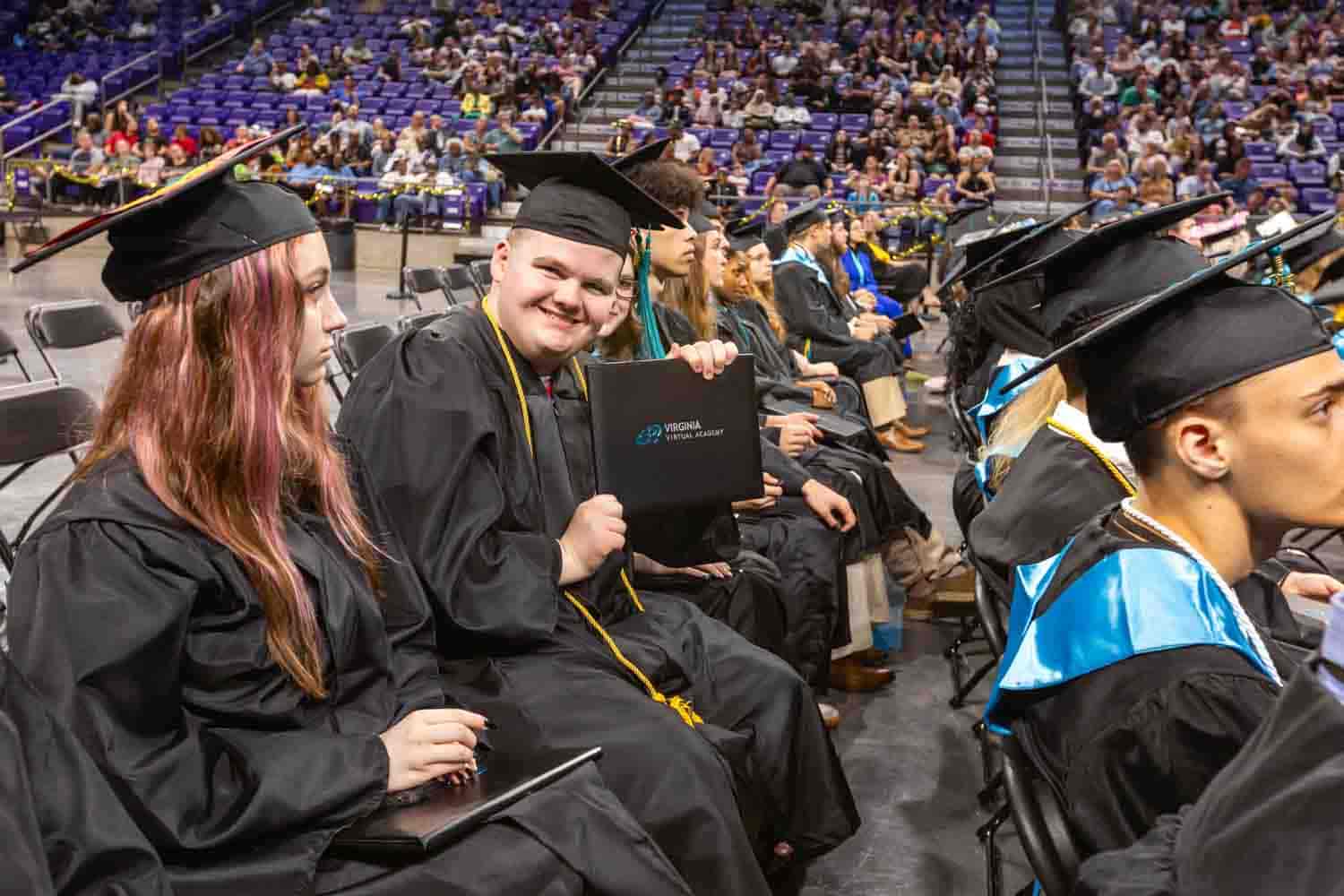 Graduates in caps and gowns sit in rows during a commencement ceremony. A smiling graduate holds a diploma, surrounded by a cheering audience.