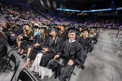 Graduates in black gowns and caps sit on chairs in a crowded auditorium, smiling and excited. An LED screen above reads "Congratulations Graduates!"