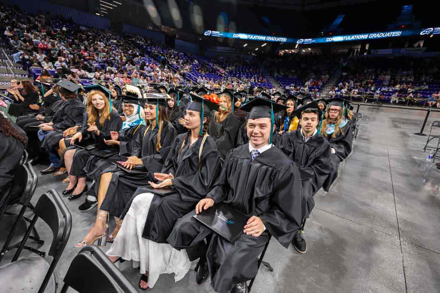 Graduates in black gowns and caps sit on chairs in a crowded auditorium, smiling and excited. An LED screen above reads "Congratulations Graduates!"
