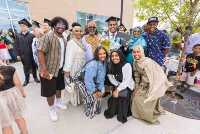 A joyful group of people, including a graduate in a cap and gown holding a diploma, celebrate outdoors. They share smiles and hugs, exuding happiness.