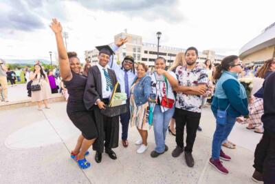A group celebrating at a graduation gathering, featuring a smiling graduate in a cap and gown holding a gift basket. The mood is joyful and festive.