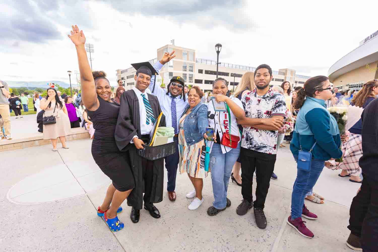 A group celebrating at a graduation gathering, featuring a smiling graduate in a cap and gown holding a gift basket. The mood is joyful and festive.