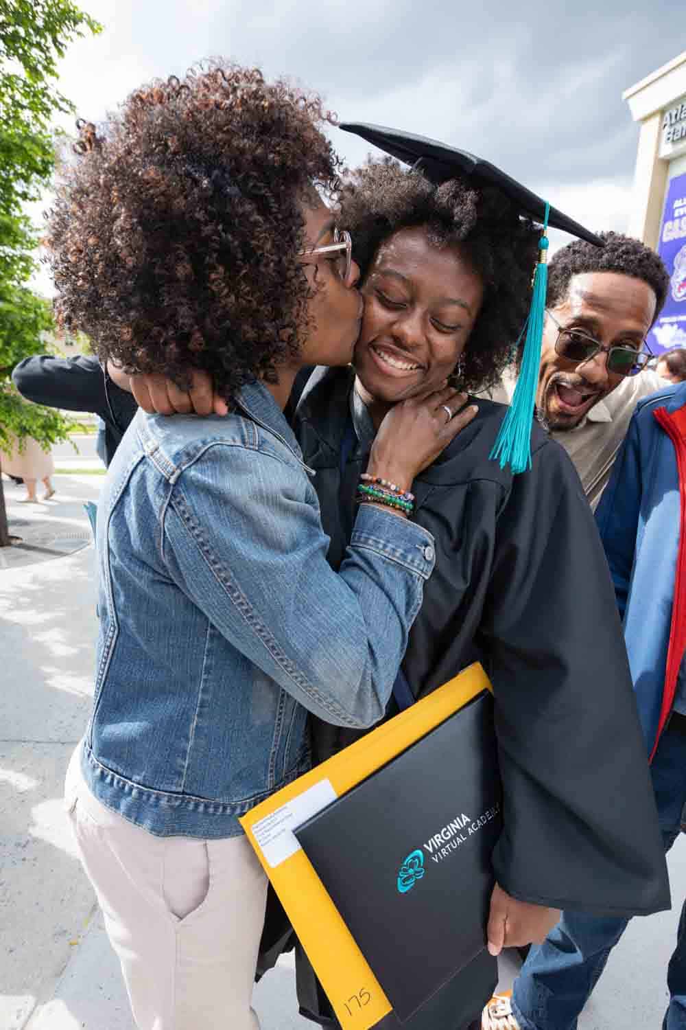 A graduate in a cap and gown is lovingly kissed on the cheek by a family member wearing a denim jacket. Another family member looks on happily. The scene conveys joy and pride.