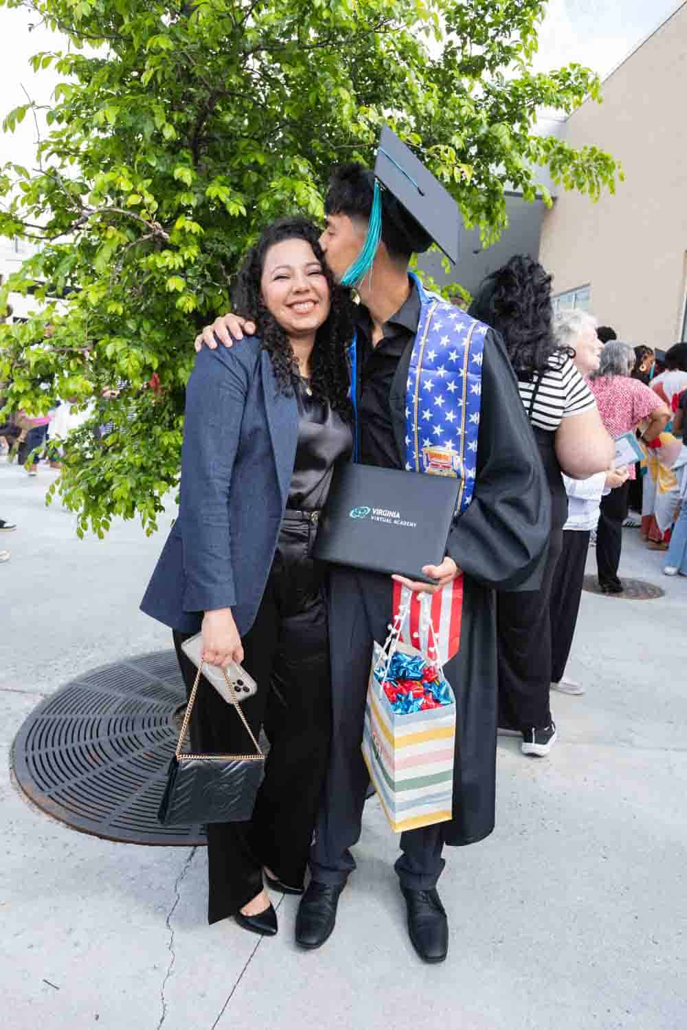 A joyful graduate in a cap and gown kisses a woman's cheek under a tree. He holds a diploma and gift bag, celebrating achievement and happiness.