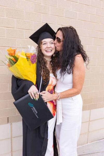 A joyful graduate in cap and gown holds flowers and a diploma, beaming as a smiling woman in white kisses her cheek against a brick wall backdrop.